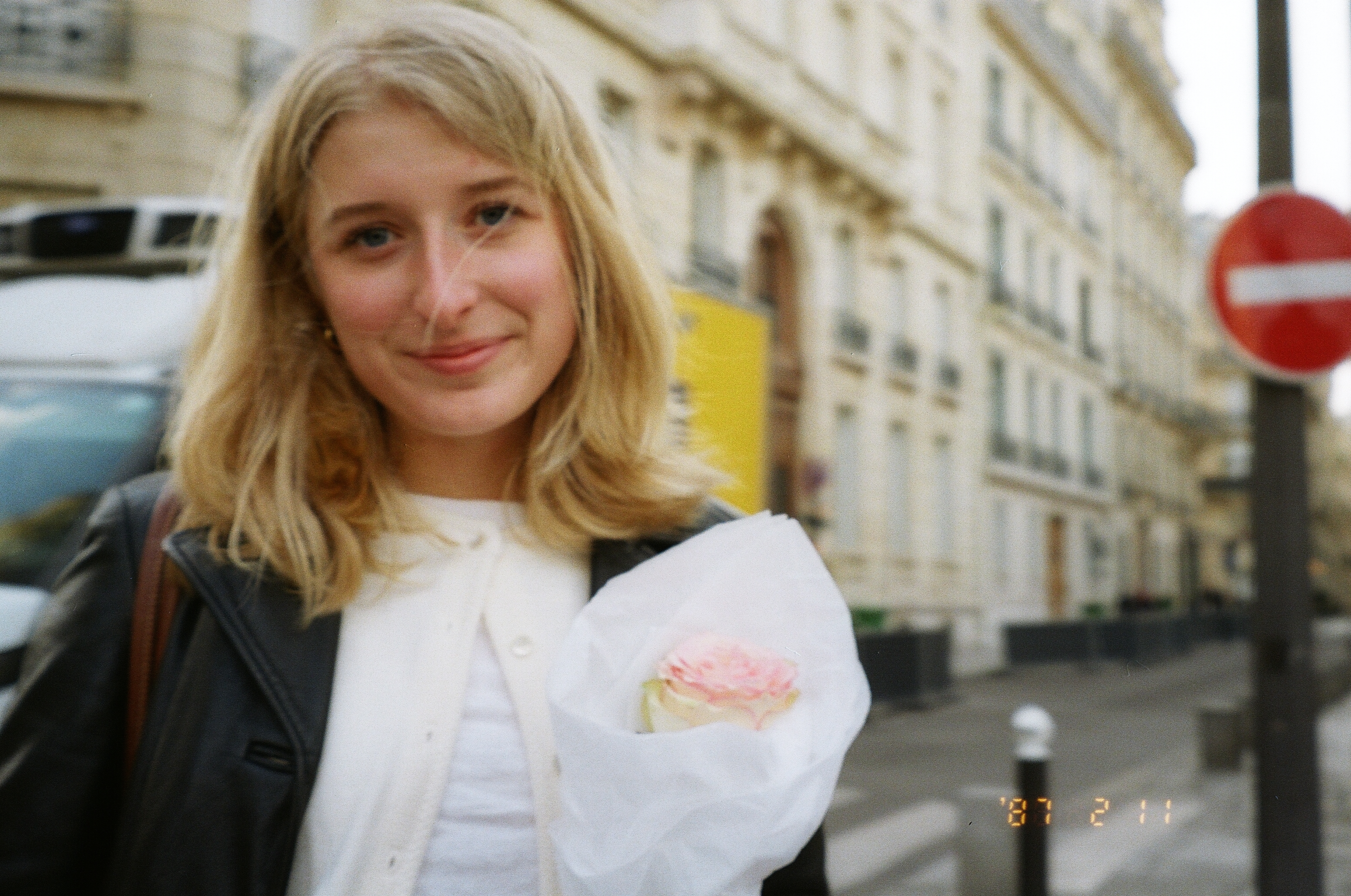 This photo depicts a 20 year old girl with blonde hair softly smiling on street while holding a light pink rose. She is standing on a street in paris, and you can see beautiful buildings behind her and a car going past. She is wearing a cream colored cardigan with a black leather jacket over top.