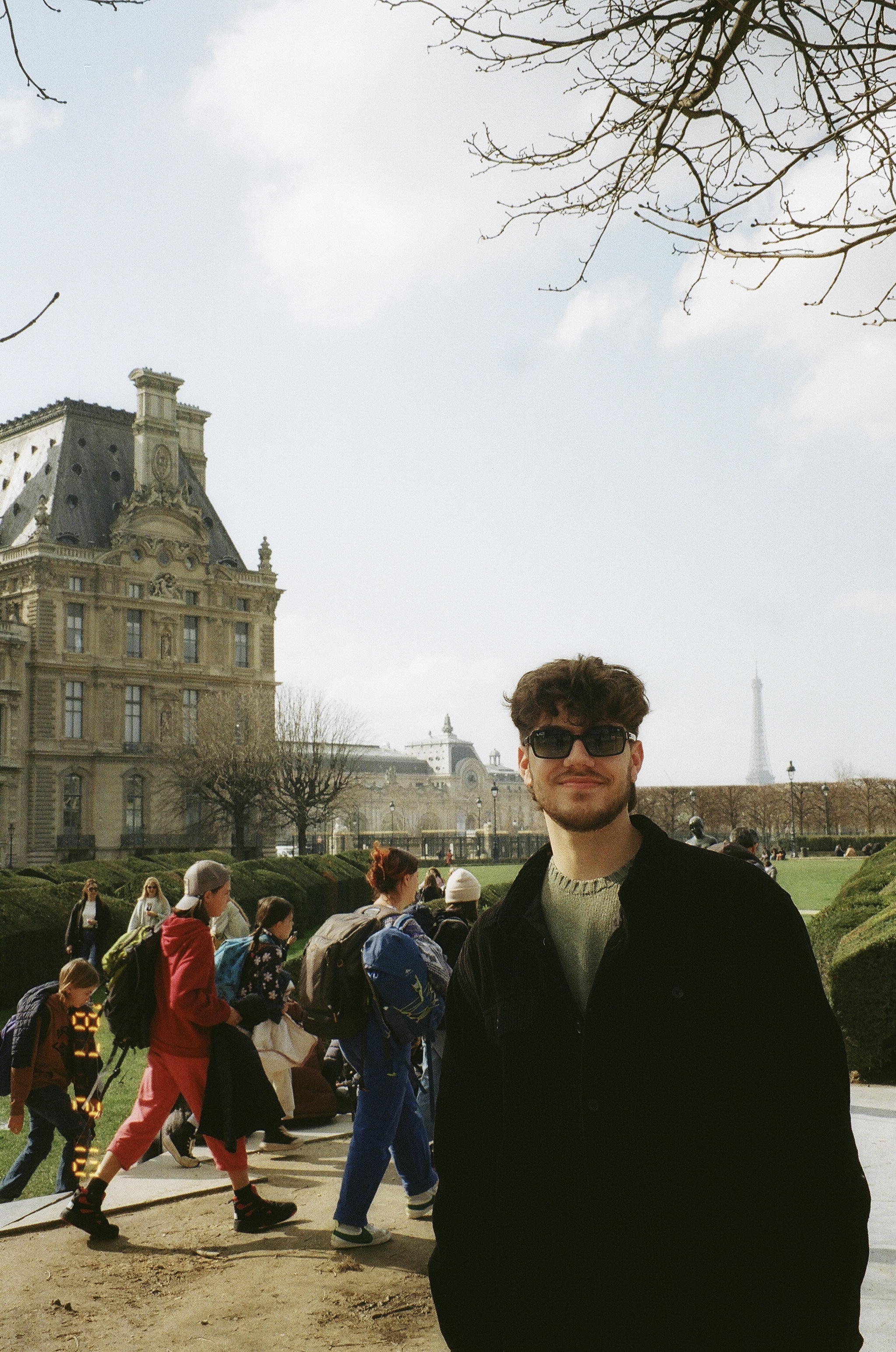 This is a photo of a young man with brown hair standing in front of the tulieres garden in Paris, with the Eiffel Tower in the distance. He is wearing a green ralph lauren sweater with black sunglasses on.