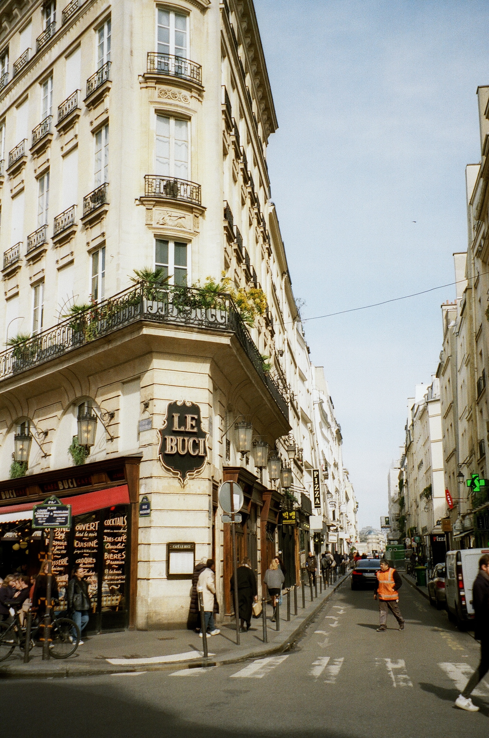 The image depicts a cafe on a corner of a cobblestone street in paris. The cafe has a bright red awning with a script logo. the street past it has many european style buildings.