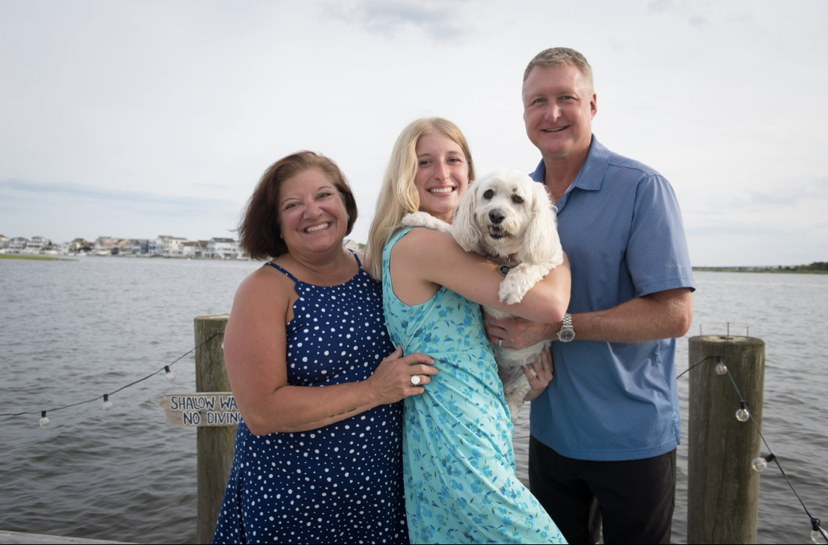 this image depicts a family of three and their white fluffy dog. They are standing on a wooden dock with an open bay behind them. In the distance you can see some other houses and trees on an island. on the left side of the image is a 55 year old woman with brown hair and brown eyes. She is hugging the girl in the middle who has blonde hair and blue eyes and is wearing a teal dress with dark blue flowers. She is holding their white dog. Next to her on the right is a 55 year old man with blonde hair and blue eyes wearing a blue shirt and he is hugging the girl in the middle from the right side