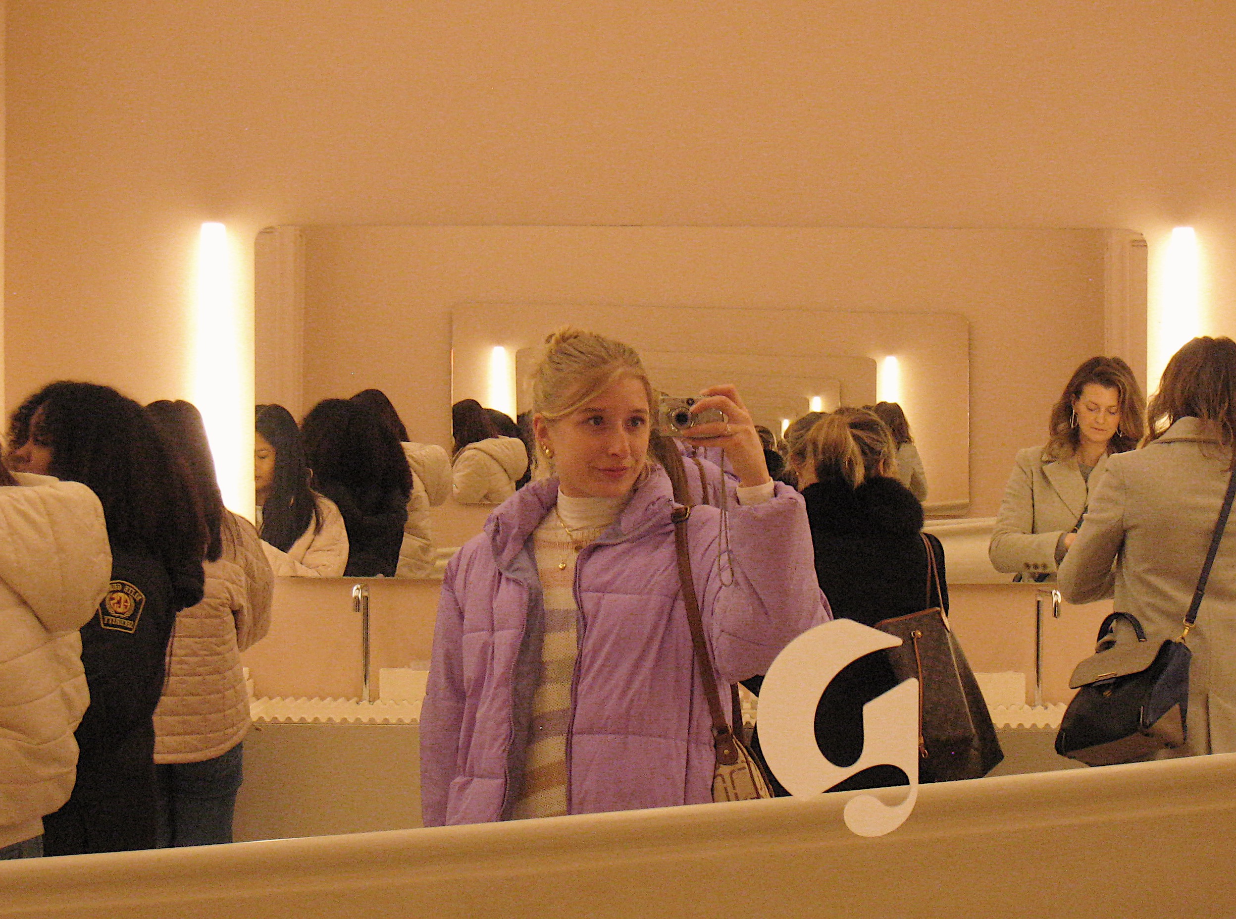 This image depicts a mirror selfie of a 20 year old girl with blonde hair up in a bun, wearing a big purple puffy coat. She is holding a digital camera and posing in the mirror of a makeup store. The walls are all a cream ish beige color.