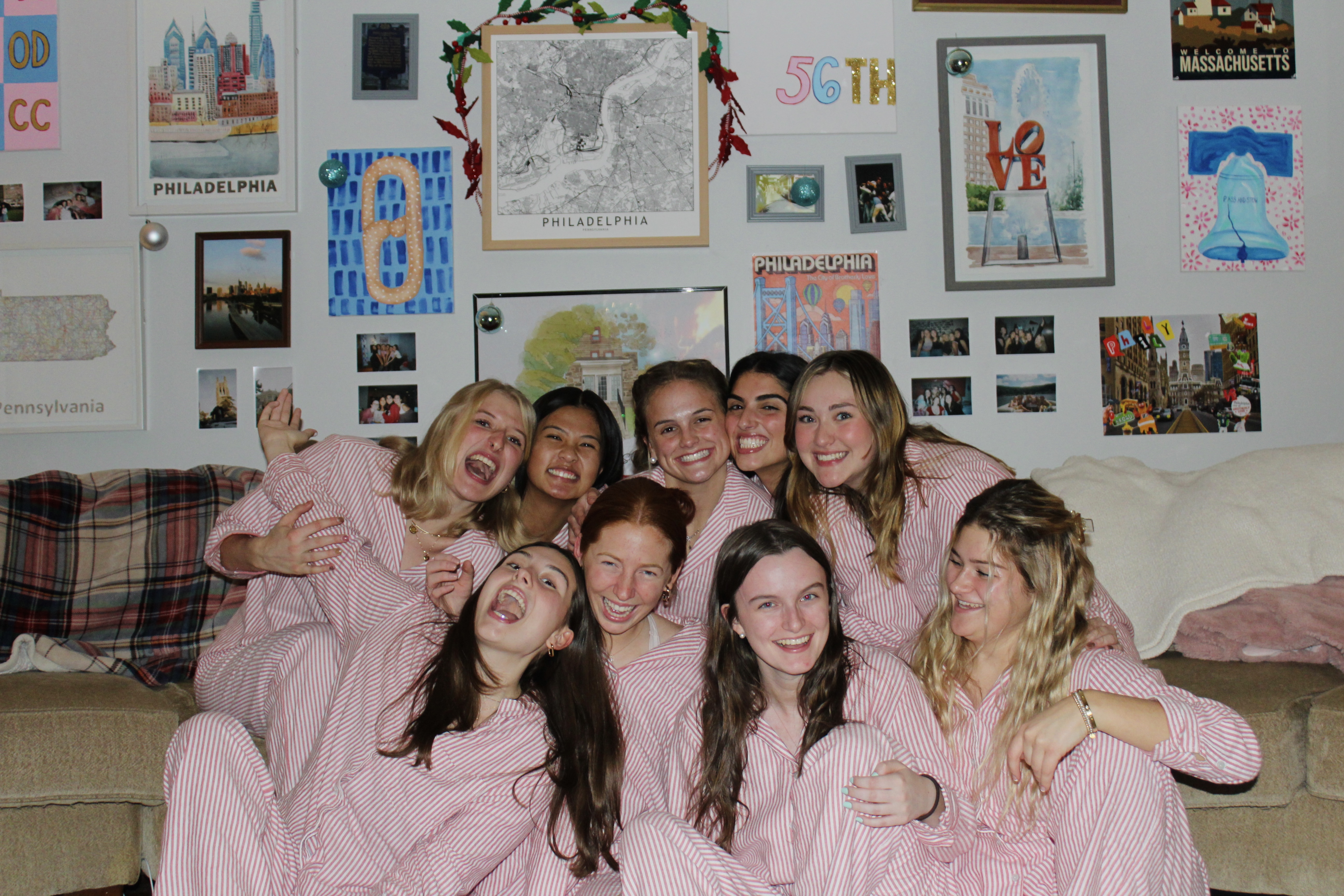 This image depicts a group of 9 girls all wearing striped christmas pajamas sitting on a big beige couch. The wall behind them has a variety of artworks and photos in frames.