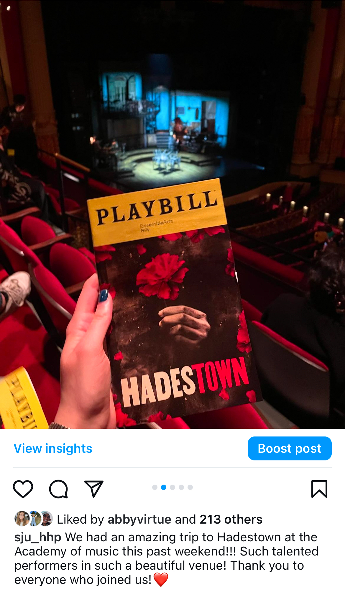 This image is a recap post from a trip to see Hadestown the musical in Philly. The center of the image has a hand holding the playbill for the musical which has a red flower in the center with the title of the show above. Behind the playbill is the audience below, and the stage behind it.